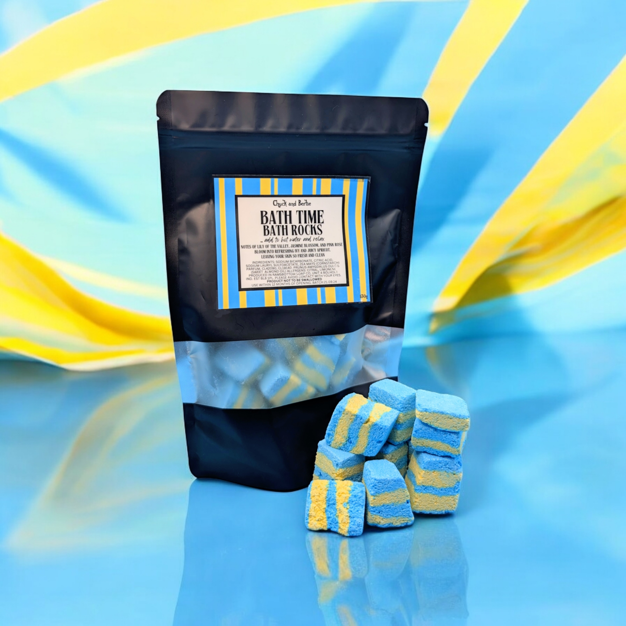 STRIKING BLUE AND YELLOW CRINKLED BACKGROUND, WITH A LARGE BLACK BAG FILLED WITH YELLOW AND BLUE PIECES OF 'BATH TIME' BATH ROCKS INSIDE AND SOME ARE POSITIONED OUTSIDE THE BAG, ON A SHINY BLUE SURFACE