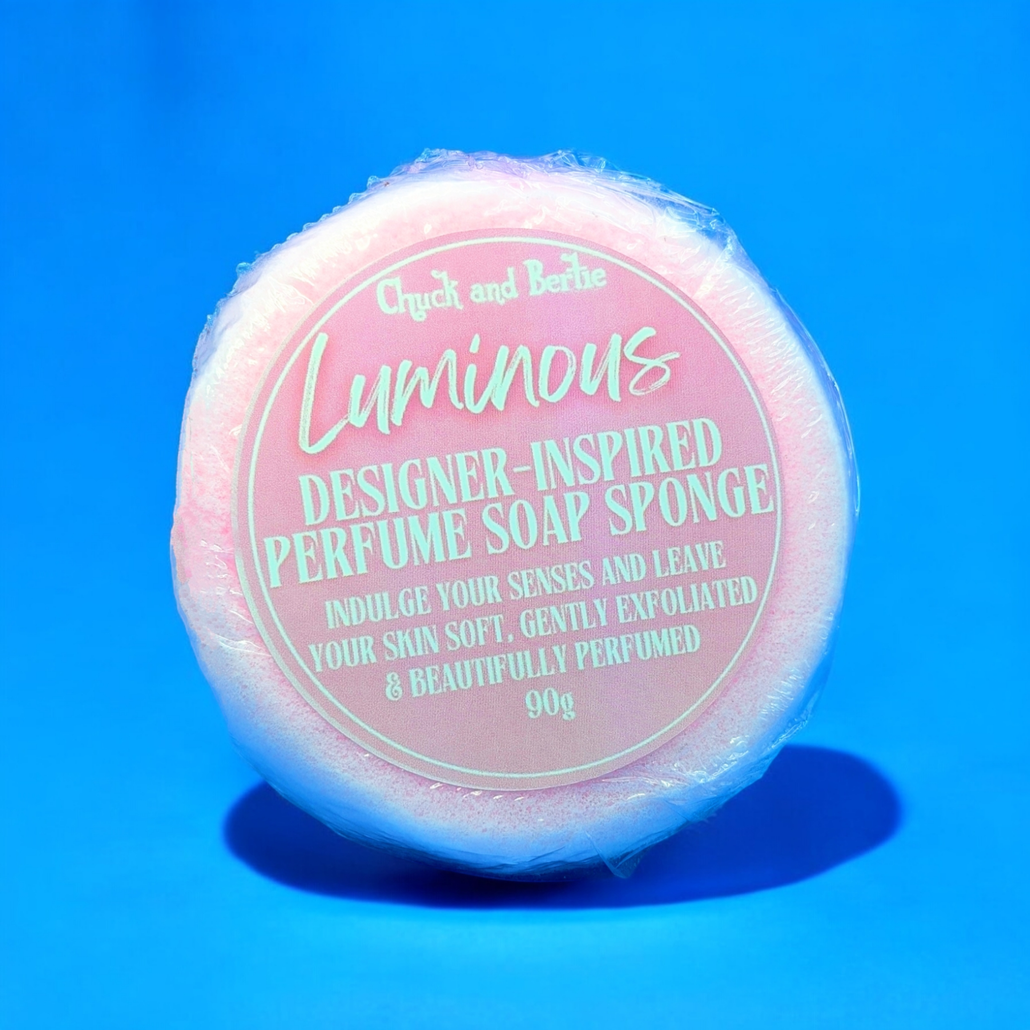 Round light pink coloured sponge in cellophane wrap with a pink and white simple and cleanly designed sticker that says "Indulge your senses and leave your skin soft, gently exfoliated and beautifully perfumed. 90 grams in weight." The background is the same shade of pink