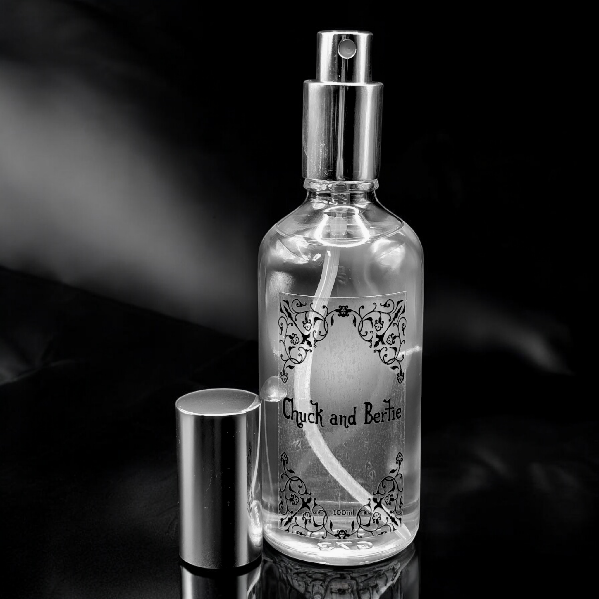 A Chuck and Bertie 100ml Fragrance Bottle with its sliver lid stood next to the bottle.  The bottle is clear shiny glass with a see-through sticker with black font